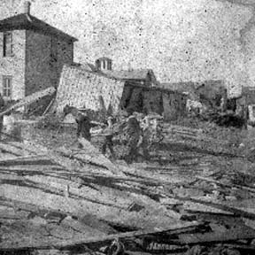 Photograph of workers carrying body past debris and wrecked houses