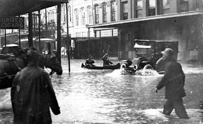 Photograph of pedestrians and automobile proceeding through flooded downtown street.� Two men in canoe