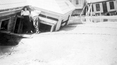 Photograph of man and woman standing in front of wrecked house partially submerged in sand 