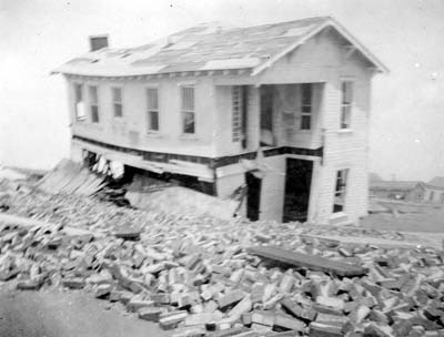 Photograph of wrecked two-story frame house viewed behind pile of bricks
