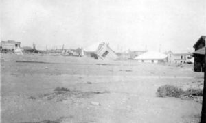 G-17713FF9.2-13 Wrecked and toppled houses partially submerged in sand
