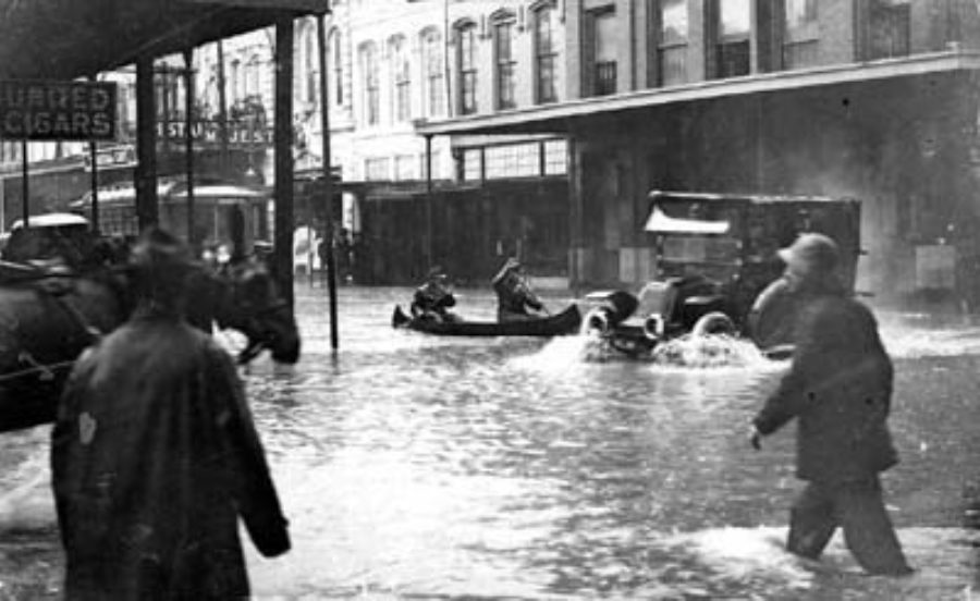 G-17713FF4.1-7 Pedestrians and automobile proceeding through flooded downtown street.